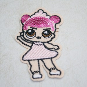 May include: Embroidered patch featuring a cartoon doll design. The doll has large brown eyes, a pink sequined hairstyle, and a pink dress. The patch is outlined in black stitching and has a beige background. The doll is in a playful pose.