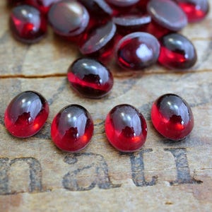Op de afbeelding: Een groep rode, ovale glazen cabochons, sommige met een lichte iriserende glans, verspreid over een houten oppervlak.