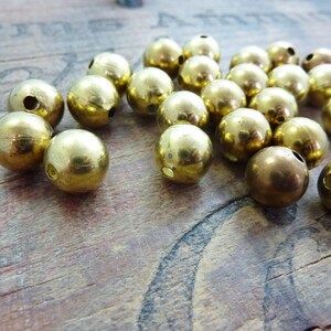 May include: A group of 20 gold-colored metal beads with a hole through the center. The beads are scattered on a wooden surface.