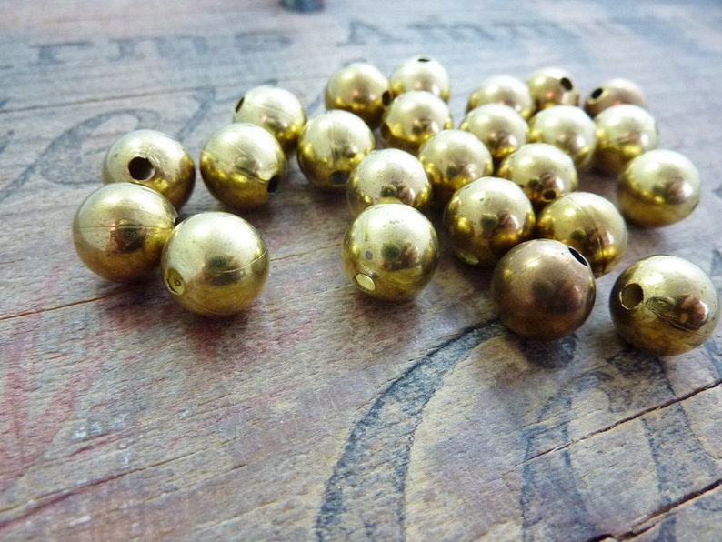 May include: A group of 20 gold-colored metal beads with a hole through the center. The beads are scattered on a wooden surface.