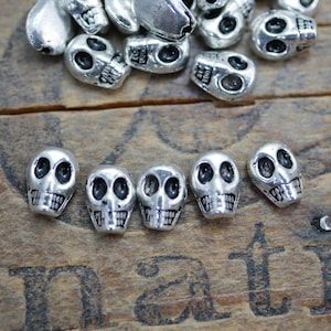 May include: A group of silver skull beads, some are laying flat and others are standing upright on a wooden surface.
