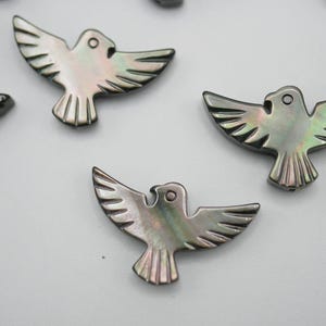 May include: Several iridescent dove-shaped pendants with open wings are displayed against a white background. The pendants have a pearlescent sheen, with details carved to resemble feathers. The dove pendants are likely for jewelry making or crafting.