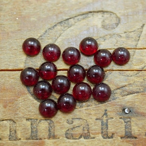 May include: A group of 15 small, round, red glass cabochons. The cabochons are arranged in a loose cluster on a wooden surface.