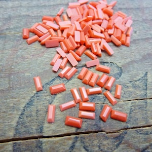 May include: A pile of small, rectangular, coral-colored beads. The beads are scattered on a wooden surface.