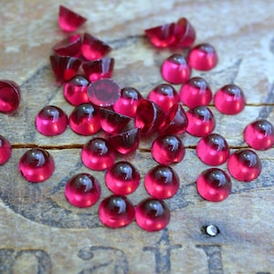 May include: A group of small, round, red glass cabochons, also known as dome-shaped beads, are scattered on a wooden surface. The cabochons are translucent and have a glossy finish.
