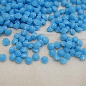May include: A close-up of a pile of light blue, round, flat-backed rhinestones with a pointed top. The rhinestones are scattered on a white surface.