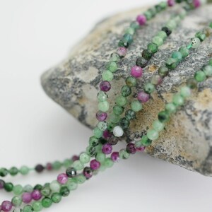 May include: A strand of green and pink gemstone beads. The beads are faceted and have a natural, irregular shape. The beads are strung on a clear thread and are displayed on a gray rock.