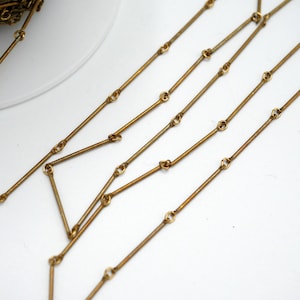May include: A close-up of a gold chain made of linked straight bars. The chain is lying on a white surface.