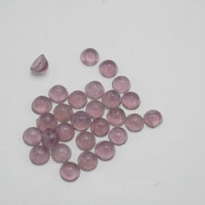 Op de afbeelding: Een groep van 25 ronde, doorschijnende, lichtpaarse glazen cabochons. De cabochons zijn verspreid over een wit oppervlak.