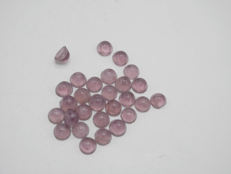 Op de afbeelding: Een groep van 25 ronde, doorschijnende, lichtpaarse glazen cabochons. De cabochons zijn verspreid over een wit oppervlak.