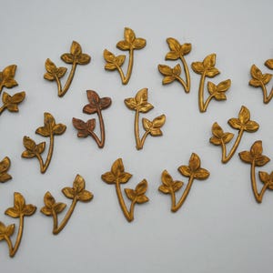 Op de afbeelding: Een verzameling van 19 goudkleurige metalen bladversieringen. Elke versiering heeft een delicaat, gestileerd ontwerp van een bladtak met meerdere bladeren.