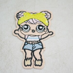 May include: Embroidered patch featuring a cartoon doll with large eyes, wearing a yellow sequined headband, a striped top, and blue denim shorts. The patch has a light tan border and is designed for application on clothing or accessories.
