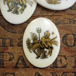 May include: White oval ceramic tile with a gold illustration of the astrological sign Aquarius. The illustration depicts a mythical creature with a fish tail and human torso holding a pitcher of water.