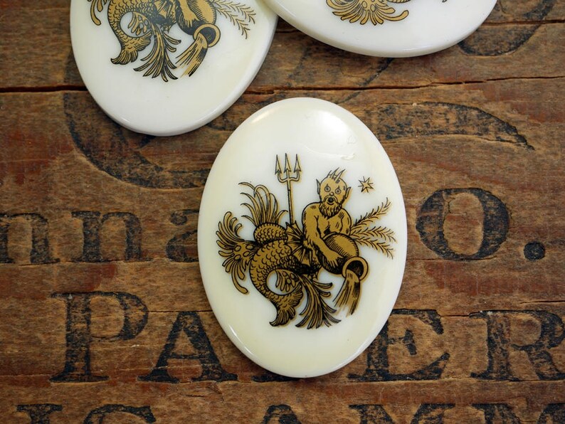 May include: White oval ceramic tile with a gold illustration of the astrological sign Aquarius. The illustration depicts a mythical creature with a fish tail and human torso holding a pitcher of water.