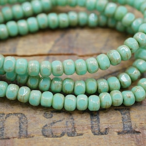 May include: A strand of turquoise blue glass seed beads with a slightly distressed finish. The beads are small and uniform in size, and they are strung together on a clear thread.