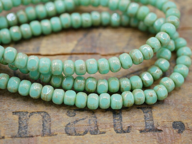 May include: A strand of turquoise blue glass seed beads with a slightly distressed finish. The beads are small and uniform in size, and they are strung together on a clear thread.