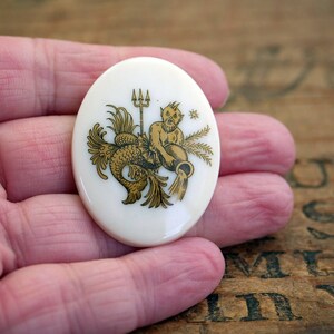 May include: A white ceramic oval pendant with a gold design of a mermaid with a trident and a crown. The pendant is on a hand.