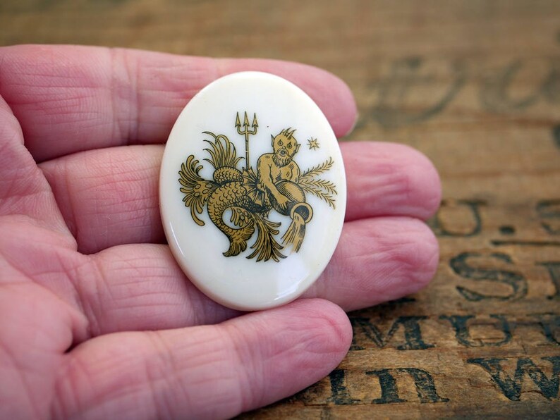 May include: A white ceramic oval pendant with a gold design of a mermaid with a trident and a crown. The pendant is on a hand.
