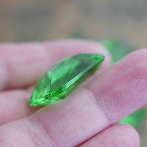 May include: A bright green faceted gemstone, cut in a rectangular shape, is held in a hand.