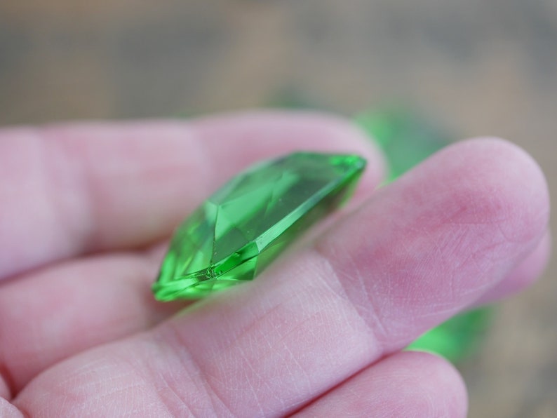 May include: A bright green faceted gemstone, cut in a rectangular shape, is held in a hand.
