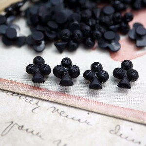 May include: Four black plastic buttons shaped like a clover, or club, from a deck of playing cards. The buttons are on a white surface with black writing.