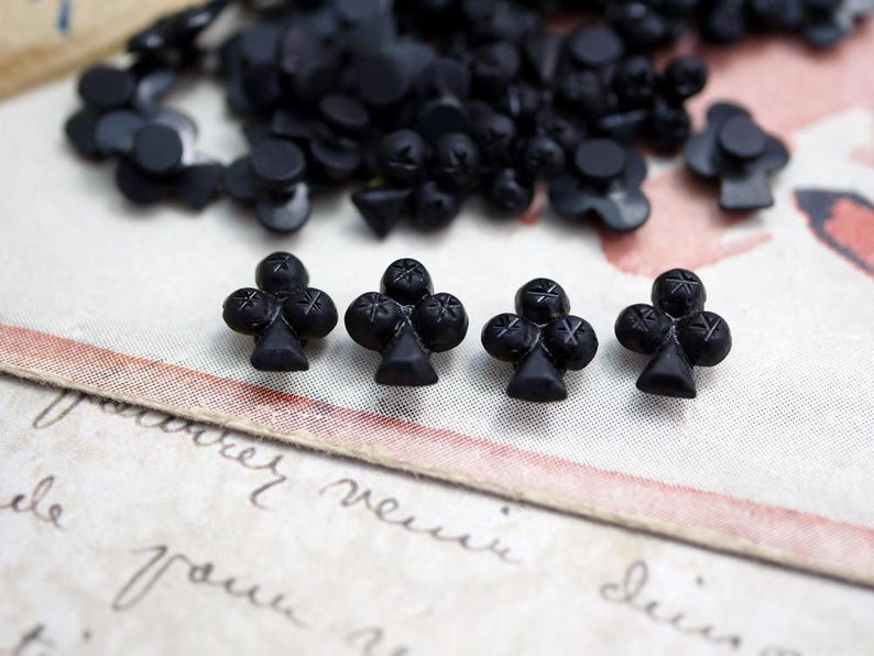 May include: Four black plastic buttons shaped like a clover, or club, from a deck of playing cards. The buttons are on a white surface with black writing.