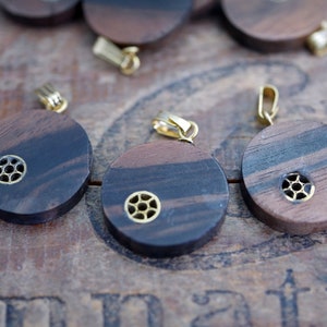 May include: Several round wooden pendants with a dark brown and black striped pattern. Each pendant has a small, gold-colored metal accent and a gold-colored bail for hanging. The pendants are displayed on a wooden surface.