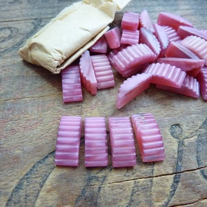 May include: A pile of pink iridescent glass tiles with a scalloped edge. The tiles are arranged in a random pattern on a wooden surface.