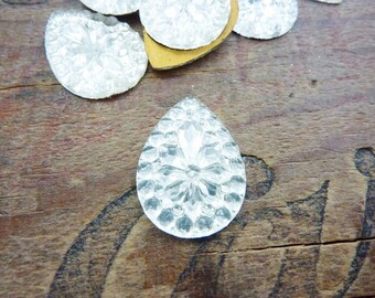May include: A clear glass teardrop-shaped embellishment with a raised, textured design. The embellishment has a faceted, crystal-like appearance.