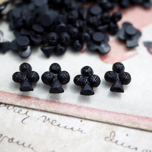 May include: Four black glass buttons shaped like a clover leaf. The buttons are on a white surface with a pink and white background.