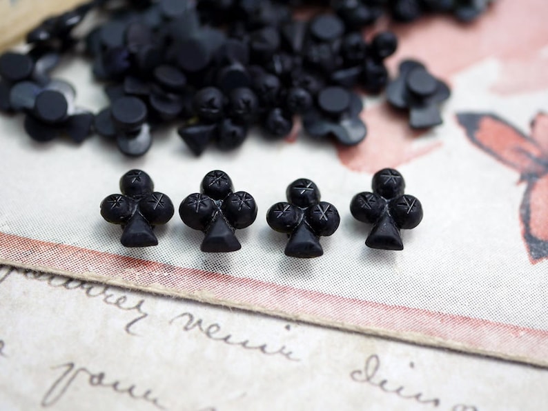 May include: Four black glass buttons shaped like a clover leaf. The buttons are on a white surface with a pink and white background.