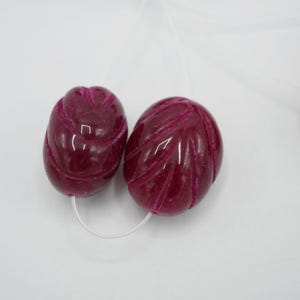 May include: Two oval-shaped, deep magenta beads with carved leaf patterns are strung on a white cord. The beads have a glossy finish and are set against a white background. These beads could be used for jewelry making.
