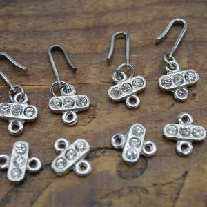 May include: Six silver-toned metal charms with clear rhinestone accents. Each charm features a cross-shaped design with three rhinestones in a row. The charms have a loop at the top for attaching to jewelry or other crafts.