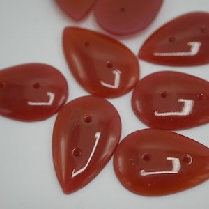 May include: A collection of teardrop-shaped, translucent red buttons. Each button has two small holes and a curved, reflective design. The buttons are scattered on a white surface, showcasing their glossy finish.