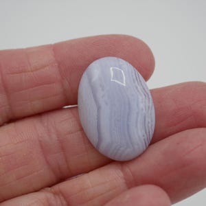 May include: A polished, oval-shaped blue lace agate gemstone. The stone features a pale blue color with swirling, banded patterns. The gemstone is held in a hand against a white background, showcasing its natural beauty.