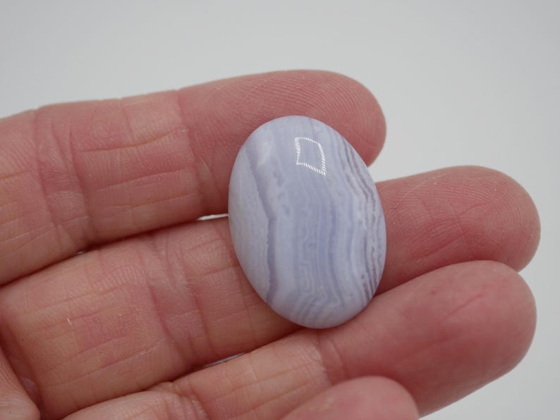 May include: A polished, oval-shaped blue lace agate gemstone. The stone features a pale blue color with swirling, banded patterns. The gemstone is held in a hand against a white background, showcasing its natural beauty.