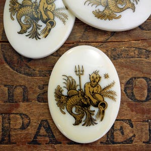 May include: White oval ceramic tile with a gold illustration of the astrological sign Aquarius. The illustration depicts a mythical creature with a human torso and a fish tail holding a pitcher of water.