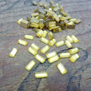 May include: A collection of small, rectangular, golden-yellow rhinestones scattered on a weathered, brown wooden surface. The rhinestones have a faceted appearance, catching the light and creating a sparkling effect. Ideal for craft projects.