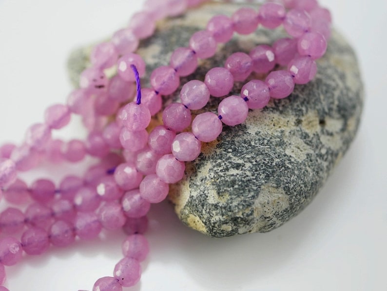May include: A strand of pink faceted gemstone beads. The beads are strung on a purple thread and are arranged in a loose pile on a grey rock.