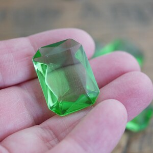 May include: A close-up of a bright green faceted glass gem. The gem is shaped like a rectangle with rounded corners and has a high polish.