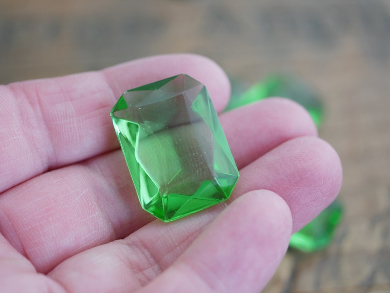 May include: A close-up of a bright green faceted glass gem. The gem is shaped like a rectangle with rounded corners and has a high polish.