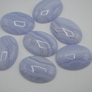 May include: Seven oval-shaped blue lace agate gemstones. Each stone has a polished, smooth surface with swirling patterns of light and dark blue. The gemstones are arranged on a white surface, showcasing their natural beauty.
