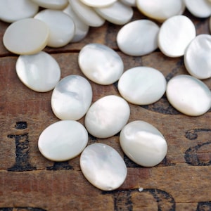 May include: A collection of oval-shaped, white mother of pearl cabochons. The cabochons are scattered on a wooden surface.