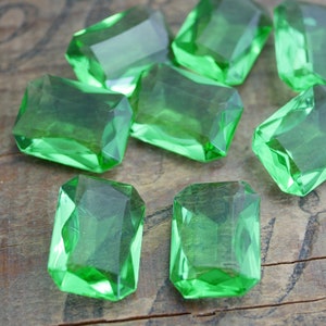 May include: A group of eight emerald green, faceted, rectangular glass gems on a wooden surface.