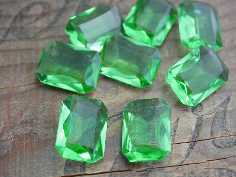 May include: A group of eight emerald green, faceted, rectangular glass gems on a wooden surface.