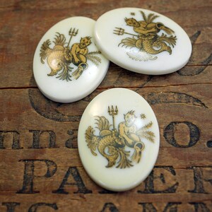 May include: Three white ceramic oval-shaped buttons with gold painted designs of the astrological sign Aquarius. Each button features a different depiction of the water bearer.