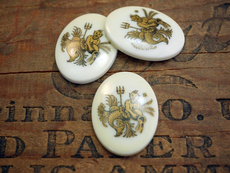 May include: Three white ceramic oval-shaped buttons with gold painted designs of the astrological sign Aquarius. Each button features a different depiction of the water bearer.
