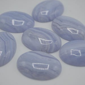 May include: Seven oval-shaped, polished blue lace agate gemstones. Each stone displays a pale blue hue with swirling, banded patterns. The gemstones are arranged on a white surface, showcasing their smooth, reflective surfaces.