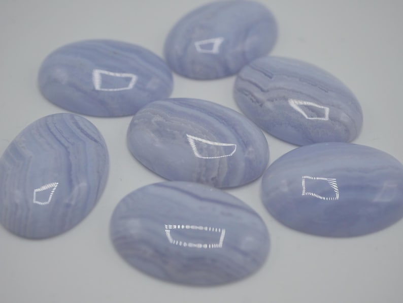 May include: Seven oval-shaped, polished blue lace agate gemstones. Each stone displays a pale blue hue with swirling, banded patterns. The gemstones are arranged on a white surface, showcasing their smooth, reflective surfaces.