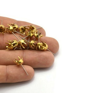May include: A close-up of a hand holding a pile of gold-coloured metal crown-shaped settings with prongs. The settings are attached to small, thin metal pins.
